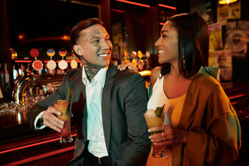 A young and diverse couple shares drinks and smiles, celebrating their relationship in a lively bar.