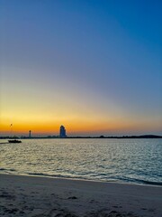 Sunset at beach. Boat near shore. Blue and orange sky with scattered clouds. Tower and tall building on horizon. Gentle waves lap against the sandy shore. A serene and picturesque coastal scene.