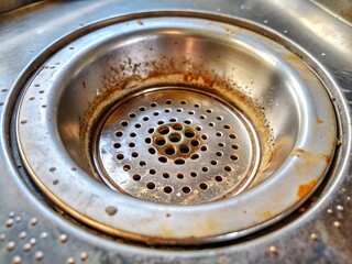 Close-up view of a kitchen sink drain reveals layers of rust and grime, reflecting years of use in a cozy, rustic kitchen. The worn metal tells a story of daily meals and gatherings