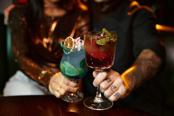 Tattooed couple clinks colorful drinks together, embracing a fun evening at a lively bar.