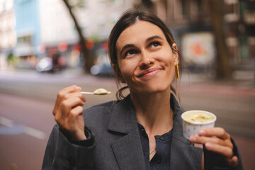 Beautiful smiling woman eating traditional ice cream in paper cup on the street. Happy dreaming woman enjoy ice cream in grey jacket.