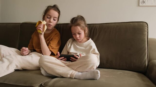 Two young sisters relax on an olive green sofa. One enjoys a banana, while the other is engrossed in a smartphone.