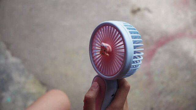 A hand is holding a portable fan and turn on then pointing it at himself. Presses the power button on a portable portable fan at summer heat