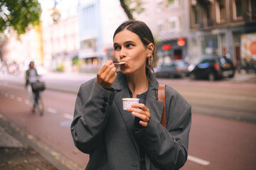 Beautiful smiling woman eating traditional ice cream in paper cup on the street. Happy dreaming woman enjoy ice cream in grey jacket.