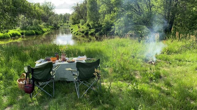 Set picnic table with food and a fire on the bank by the river. Panoramic view of the river. Atmospheric futage of rest in nature, solitude, wild vacation, no people