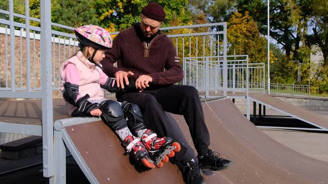 A father helps his little daughter put on her roller skating gear at a skate park. They re sitting on a ramp, a sunny autumn day