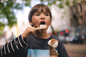 Little brunette boy eat ice cream in waffle corn with spoon. Happy childhood. Boy walk outdoor in the city street. 