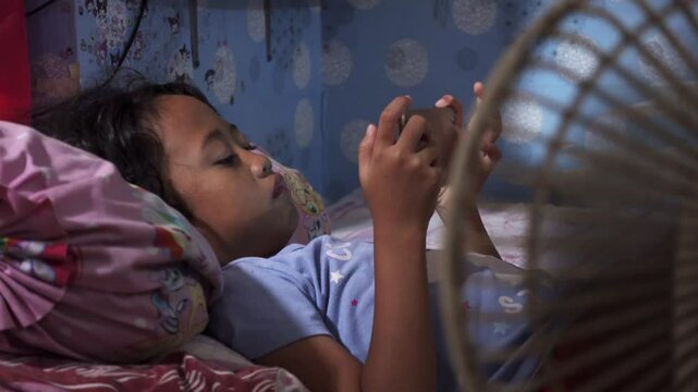 Girl playing smartphone on hot summer say while bedridden near fan. Cool wind from electric fan at summer heat.
