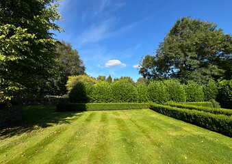 A well-maintained garden features neatly trimmed hedges and a lush green lawn under a bright blue sky. Tall trees border the space, creating a tranquil and inviting atmosphere near, Riddlesden, UK