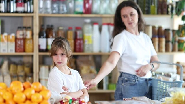 Angry child is standing in grocery store, capricious and sulky silence. Girl makes faces and ignores conversation with her mother, parent screams and is indignant in background