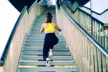 Back view of caucasian female athlete with slim figure doing squatting exercises on stairs during workout, woman in sport activewear reaching fitness goals training legs muscles and vitality outdoors