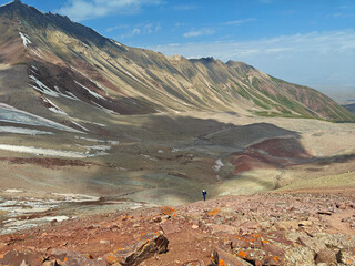 A lone hiker climbs vibrant red and green rocks beneath a clear blue sky in a serene mountain...