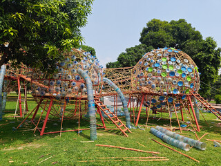 Outdoor installation made from upcycled plastic bottles and materials, featuring dome-shaped structures connected with tubes and surrounded by greenery in a park setting under bright sunlight