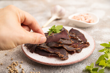 Armenian Basturma dried meat with hand on plate with pepper and herbs on brown concrete. Side view, selective focus
