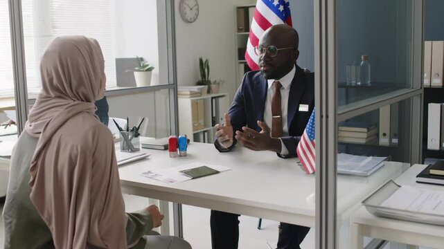 Arc shot of Muslim woman in hijab applying for US visa, having interview with Black male visa officer in embassy