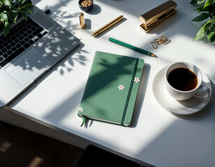 a green notebook, a cup of coffee and a laptop on a clean white desk with plants arround it