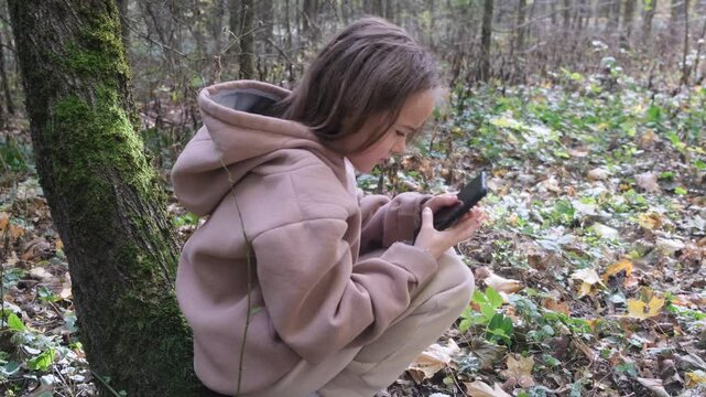 A young girl sits crouched in a forest, looking at her smartphone with a look of distress. The phone appears to be off or have a dead battery, adding to her apparent worry and isolation in the woodlan