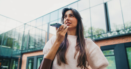 Beautiful Caucasian girl recording voice message on smartphone. Talking to microphone while holding mobile device horizontally with one hand. Thinking what to say to friend or relative.