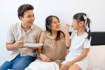 Fototapeta premium Asian father hold dish of food and give to little girl with pregnant mother stay on bed in concept of happiness family in their house.