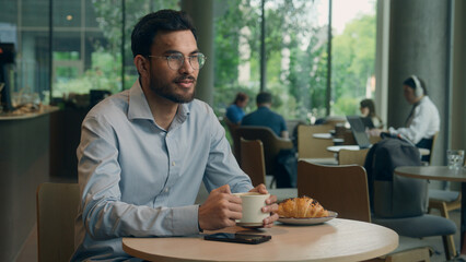 Indian Arabian guy hungry man male businessman visitor take away order eat croissant in cafe restaurant bakery cafeteria eating food enjoy taste drink coffee breakfast business lunch healthy nutrition