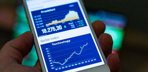 Hand holds a smartphone which displays a rising technology stock index. Close-up smartphone display with various stock indice. Business, investment, trading, online brokerage.