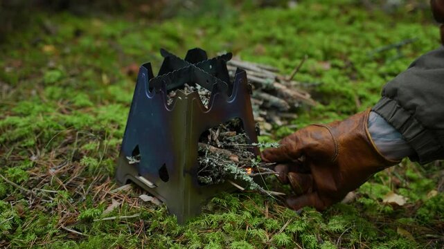 View of a hand in a leather glove lighting a fire with a match in a portable camping stove. Starting a fire while hiking. Camping wood stove for cooking food. Close-up