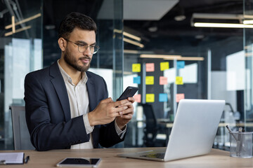 A focused businessman using a smartphone while sitting at a desk with a laptop in a modern office...