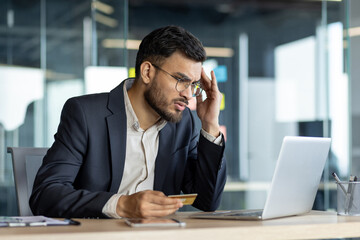 A businessman expresses stress as he holds a credit card while working on a laptop. The office...