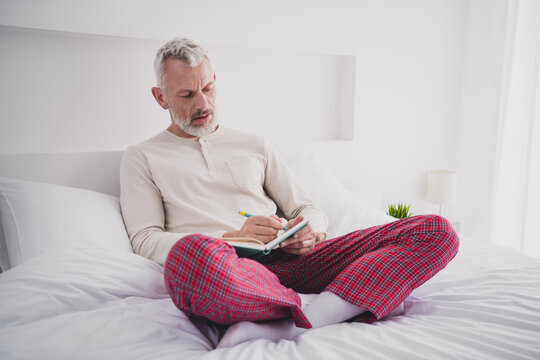 Mature man enjoying a relaxing morning in pajamas, writing in a journal while sitting comfortably on a bed