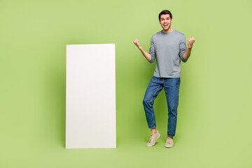 Full body photo of overjoyed delighted man raise fists in victory next to long white board promo isolated on green color background