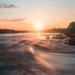 Golden sunlight dances on the rushing waters of the Guaviare River, as day transitions to night in the heart of Colombia's lush Amazon forest, creating a tranquil and enchanting atmosphere.
