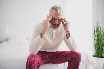 Mature man experiencing morning head pain while sitting on bed in bright bedroom