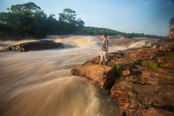 Standing on rocky terrain, an individual gazes at the powerful currents of the Guaviare River. Bustling waters reflect the soft glow of the early morning sun against lush greenery