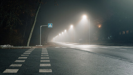 Foggy night scene with an illuminated pedestrian crossing and a row of glowing streetlights fading into the mist.