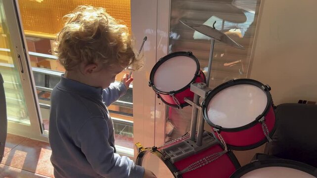 Young boy enthusiastically plays a bright red drum set in a sunny room, filling the space with joyful music and energy