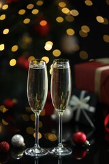 Delicious champagne in glasses and Christmas decor on black mirror surface against blurred lights, closeup