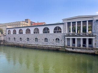 Ljubljana, Slovenia - June 28, 2024: Covered market along the Ljubljanica river with Institute of Labor Law at the University building in back under blue sky