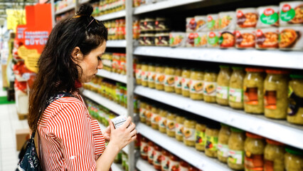 Woman carefully examining a product in a supermarket aisle filled with jars of preserved foods.