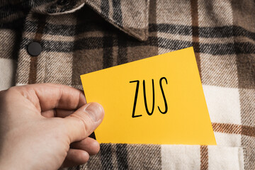 Yellow card with a handwritten inscription "ZUS", a shaft in the hand, protruding from a brown plaid shirt (selective focus)