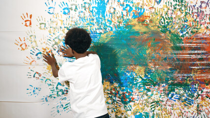 Back view of happy african boy painted the stained wall with colorful hand while wearing casual white shirt in art lesson.Smart student use hand print to make creative artwork. Education. Edification. © InfiniteFlow
