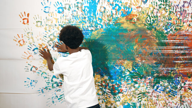 Back view of happy african boy painted the stained wall with colorful hand while wearing casual white shirt in art lesson.Smart student use hand print to make creative artwork. Education. Edification.
