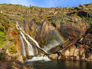 A scenic waterfall cascades down a rocky cliff into a pool below. Greenery and vegetation are scattered on the rocks. A rainbow appears in the mist, adding color to the bright, sunny day.