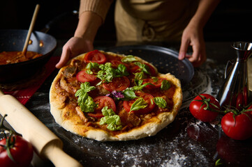 Person preparing a fresh pizza with tomatoes and basil in a kitchen