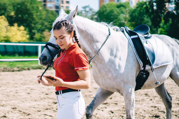 Caucasian horsewoman using 4g wireless internet during walk in paddock of country club, active...