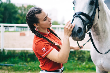 Caucasian professional horsebacker take care about friend animal standing outdoors at nature environment and talking to horse, young brunette girl spending day for practicing equestrian sport