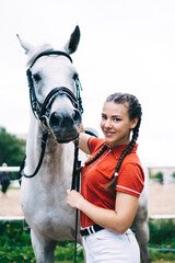 Obraz premium Half length portrait of Caucasian jockey girl posing during dressage competition at nature, female horseback rider smiling at camera during hobby equestrian with white champion stallion animal
