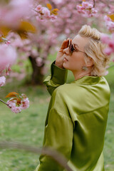 Fototapeta premium A blonde woman in green outfit poses surrounded by cherry blossoms outdoors