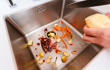 Peeling potatoes over a kitchen sink filled with food scraps and running water.