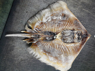 A dried ray fish, flat and wide, with its fins spread out, shows a smooth texture and light brown color.