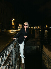 A stylish woman is confidently posing near a scenic canal at night